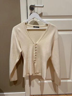 Krämvit kofta från Gina Tricot - Snygg krämvit kofta från Gina Tricot med v-ringning och knappar framtill. Långärmad och i mjukt, tunt stickat material. Perfekt att slänga över en topp för en chill look.