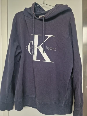 Mörkblå hoodie från Calvin Klein Jeans - Snygg mörkblå hoodie från Calvin Klein Jeans med stor vit CK-logga och text på bröstet. Klassisk modell med huva och dragsko samt ribbade muddar vid ärmslut och nederkant. Perfekt för en avslappnad och trendig stil.