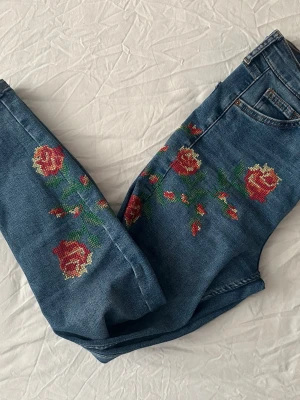 TopShop Blå jeans med broderade rosor - Snygga blå jeans med broderade röda rosor och gröna blad på båda benen. Modellen är slim fit med klassisk femficksdesign och normal midja. Perfekta för dig som vill sticka ut med coola detaljer och unik stil. Storlek W28L30