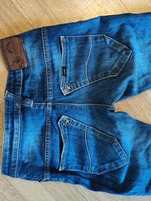 Jeans från Lee stl 25/31 - Säljer ett par jättesnygga blå jeans från Lee med coola slitningar och dragkedjor vid bensluten. Jeansen har unika bakfickor med dekorativa sömmar och Lee-logga. Klassisk femficksmodell. Lee Louis zip stl 25/31 i bra skick.