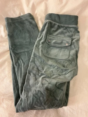 Gröna velourbyxor från Juicy Couture - Mjukisbyxor i grön velour från Juicy Couture med bred resår i midjan och dragsko. Byxorna har två stora framfickor och en bakficka med lock och tryckknappar. Liten Juicy Couture-logga broderad på ena fickan framtill. Perfekta för en chill och avslappnad stil.