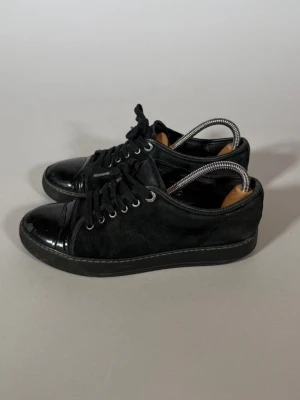 Svarta låga sneakers med lackdetaljer - Snygga svarta sneakers med både mocka och lackdetaljer på tå och häl. Skorna har svarta skosnören och metallfärgade öljetter. Perfekta för dig som gillar en stilren och modern look med lite extra edge.