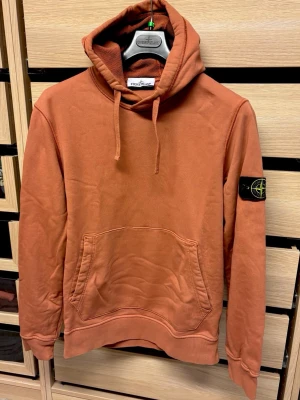 Rustbrun hoodie från Stone Island - Säljer en rustbrun hoodie från Stone Island med klassisk patch på vänster ärm. Hoodien har stor känguruficka framtill, dragsko i huvan och ribbade muddar. Perfekt för dig som gillar streetwear och vill ha något bekvämt och snyggt.