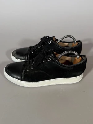 Svarta låga sneakers med vit sula - Stilrena svarta sneakers med lågt skaft och vit sula. Skorna har snörning, ovandel i mocka och läder samt klassisk siluett. Perfekta för en clean och avslappnad look.