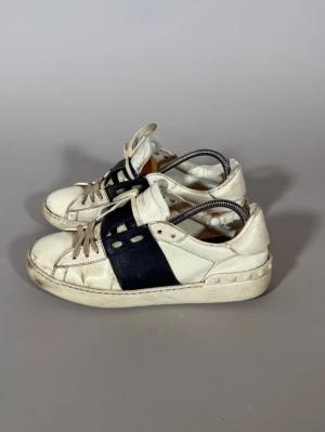 Valentino vita sneakers med svart detalj - Snygga vita sneakers från Valentino med bred svart läderdetalj på sidan och klassisk snörning. Skorna har rund tå, låg profil och diskret logga på plösen. Perfekta för dig som gillar stilrena och exklusiva sneakers.