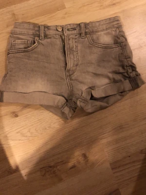 Grå jeansshorts från Kappahl - Snygga grå jeansshorts från Kappahl med klassisk femficksmodell, uppvikta benslut och dragkedja med knapp. Perfekta för sommardagar och enkla att matcha med olika toppar. Materialet är slitstarkt denim i bomull.