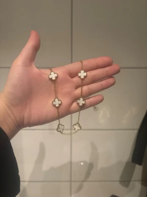 Guldhalsband med vita klöverhängen - Snyggt halsband i guldton med flera vita klöverformade hängen. Hänget har en pärlemorskimrande yta och halsbandet har en tunn kedja. Perfekt accessoar för att lyfta din outfit med en elegant touch.