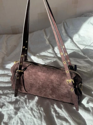 Brun mocka handväska  - Snygg handväska i brun mocka med cylinderformad design och justerbara axelremmar i läder. Väskan har guldfärgade metalldetaljer och dragkedja upptill. Perfekt för dig som gillar en stilren och trendig look.