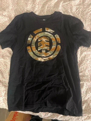 Svart Element t-shirt med camologga - Svart t-shirt från Element med stor logga i camouflagemönster på bröstet. Klassisk rund hals och korta ärmar. Perfekt för dig som gillar streetwear och skatekultur.