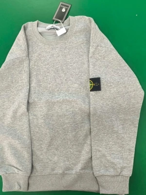 Grå sweatshirt från Stone Island - Säljer en stilren grå sweatshirt från Stone Island med klassisk rund halsringning och ribbade muddar. Tröjan har den ikoniska Stone Island-loggan på vänster ärm och är tillverkad i mjuk bomull för maximal komfort.