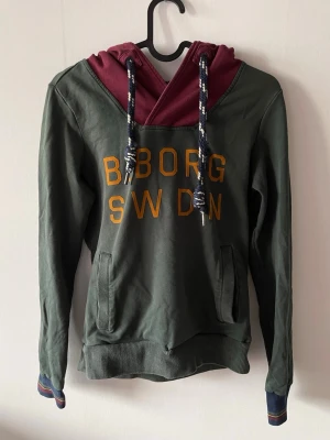 Grön hoodie från Björn Borg - Mörkgrön hoodie från Björn Borg med vinröd huva och stora gula bokstäver på bröstet. Snygga detaljer som grova snören i huvan och ribbade muddar med randig kant. Praktiska fickor framtill. 