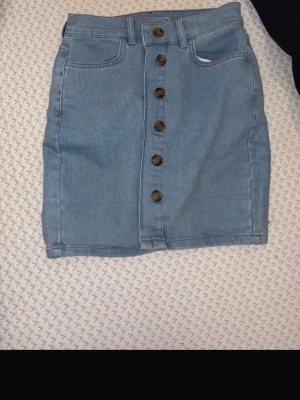 Ljusblå jeanskjol med knappar Janina - Ljusblå jeanskjol från Janina med snygg knappslå framtill. Kjolen har klassiska fickor fram och bak, och är i en kort modell som passar perfekt till vardagslooken. Tillverkad i bomull för en bekväm känsla.