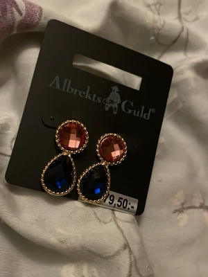 Stora statement örhängen Albrekts Guld - Snygga statement örhängen från Albrekts Guld med runda, roséguld-färgade stenar upptill och droppformade mörkblå stenar nedtill. Båda stenarna är infattade i guldfärgad ram med små pärldetaljer runt om. Perfekta för att lyfta vilken outfit som helst.