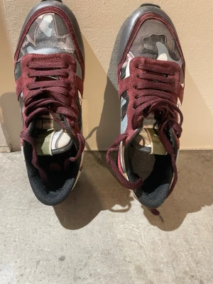Valentino Rockrunner sneakers med camomönster - Säljer ett par Valentino Rockrunner sneakers med burgundy röda detaljer och camouflagemönster i grått, svart och beige. Skorna har snörning, platt sula och inslag av mocka och skinn. Snygga nitar baktill och rund tå ger en cool vibe. Äkta✅