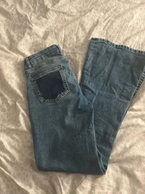 Blå utsvängda jeans med patch - Säljer ett par blå jeans med utsvängda ben och en cool mörkblå patch på bakfickan. Jeansen har klassisk femficksdesign och är gjorda i denim. Perfekta för dig som gillar retrostil och vill sticka ut lite extra. 
