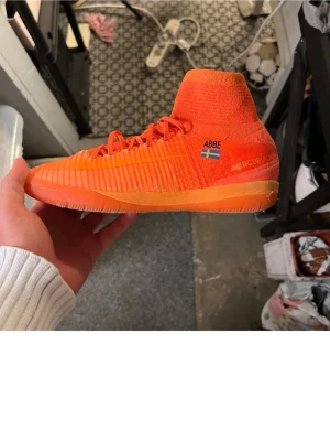 Nike Mercurial orange fotbollssko - Snygg orange Nike Mercurial fotbollssko med högt skaft och snörning. Skon har texten 'ABBE' och en svensk flagga på sidan samt Mercurial-logga. Tillverkad i syntetmaterial med ribbad struktur för bättre bollkontroll. Perfekt för dig som vill sticka ut på planen.