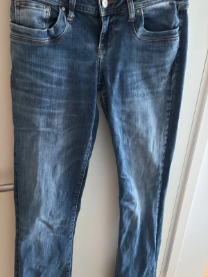 Blå raka jeans med slitningar - Säljer ett par klassiska blå jeans med bootcut och lätt slitna detaljer. Jeansen har fem fickor, knappgylf och är tillverkade i denim med en snygg tvättad look. Perfekta för en avslappnad stil. W27 L34