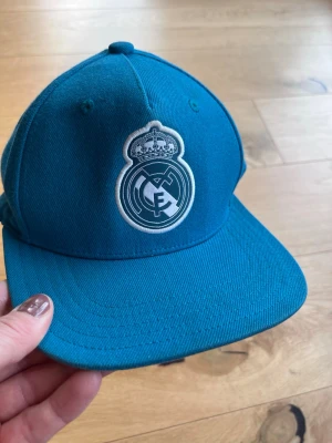 Blå Real Madrid keps junior - Snygg blå keps med Real Madrid-logga broderad framtill. Klassisk modell med böjd skärm och justerbar baksida. Perfekt för dig som vill visa vilket lag du hejar på. Materialet känns slitstarkt och kepsen har en sportig vibe.