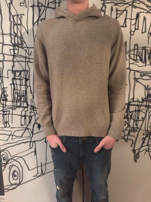 Stickad hoodie  - Säljer en stilren beige hoodie i mjuk ullmix med ribbade muddar vid ärmslut och nederkant. Klassisk passform och huva utan snören. Perfekt för dig som gillar en enkel men snygg look.