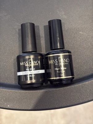 Miss Fancy Top Coat Nagellack Duo - Två professionella top coat nagellack från Miss Fancy. Ena flaskan är en No Wipe Glitter Top Coat för extra glans och glittereffekt, den andra är en High Shine Top Coat som ger en spegelblank finish. Båda flaskorna är svarta med tydliga etiketter och har klassisk rund form.