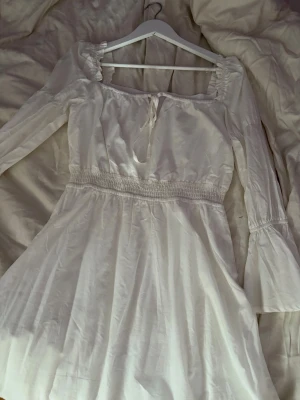 Vit kort klänning med knyt - Söt vit kortklänning med långa ärmar och smock i midjan. Klänningen har en rundad urringning med knytband och lätt volang vid axlarna. Tillverkad i ett mjukt, tunt tyg som ger en luftig känsla. Perfekt för sommarens alla tillfällen.