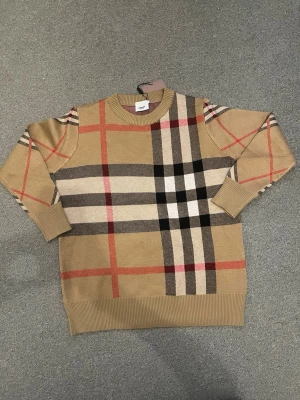 Beige rutig tröja från Burberry - Snygg beige tröja från Burberry med klassiskt rutmönster i svart, vitt och rött. Tröjan har rund halsringning, ribbade muddar och är tillverkad i mjukt stickat material. Perfekt för dig som gillar stilrena och ikoniska plagg.
