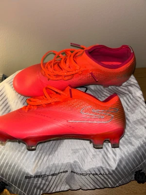 Röda fotbollsskor från Skechers - Snygga fotbollsskor från Skechers i en intensiv röd färg med orange och silvriga detaljer. Skorna har snörning, låg profil och mönstrad sula för bra grepp på planen. Ovandelen är i syntetmaterial och skon har en sportig, modern design.