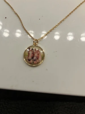 Guldhalsband med hästmotiv - Unikt halsband med tunn guldfärgad kedja och ett runt hängsmycke med motiv av två hästar. Hängsmycket har en glansig yta och ramen är i guldfärg. Perfekt för dig som älskar hästar och vill ha något personligt och sött runt halsen.