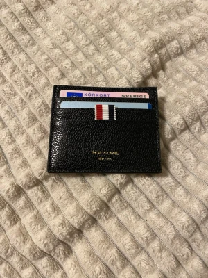 Svart kortfodral från Thom Browne New York - Snyggt svart kortfodral i präglat läder från Thom Browne. Fodralet har flera kortfack och en ikonisk randig detalj i rött, vitt och blått på framsidan. Guldtext med logga på framsidan ger en exklusiv känsla. Perfekt för dig som vill ha stilren och lyxig accessoar.