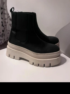 Svarta chunky Chelsea boots - Säljer ett par svarta Chelsea boots med riktigt chunky, beige platåsula. Skorna har elastiska paneler på sidorna och dragflik bak. Tillverkade i slätt material som ser ut som skinn. Perfekta för dig som vill sticka ut med din stil.