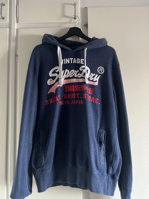 Blå Superdry hoodie med tryck - Mörkblå hoodie från Superdry med vit och röd logga och text på bröstet. Klassisk huva med vita snören, köpt för 1300kr och säljs för 450 väldigt skön o rätt tjock att ha på sig nu när de är kallt men ändå väldigt snygg, pris är ej hugget i sten och de bara att höra av sig vid funderingar🙌🙌