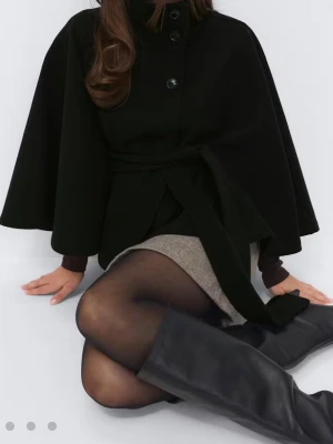 Svart cape-kappa med knytband - Stilren svart cape-kappa med knappar framtill och brett knytband i midjan. Kappan har vid siluett och hög krage. Perfekt för dig som vill ha något extra till garderoben. Materialet ser ut att vara ullblandning. Använd 1 gång men tyckte inte den passa mig så väljer att sälja där av priset. Storlek xs/s