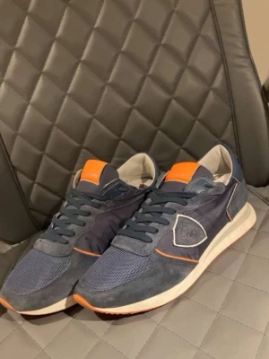 Blå sneakers från Philippe Model - Snygga sneakers i blått från Philippe Model med detaljer i orange och vitt. Ovandelen är i mocka och mesh för en sportig look. Skorna har vita och orange detaljer på sulan samt en ikonisk logga på sidan. Perfekta för dig som gillar streetstyle.