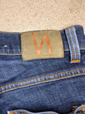 Mörkblå jeans från Nudie Jeans - Säljer ett par mörkblå jeans från Nudie Jeans med klassisk femficksmodell och snygga kontrastsömmar. Jeansen har en rak passform och är gjorda i slitstark denim. Perfekta för dig som gillar stilrena och tidlösa jeans.