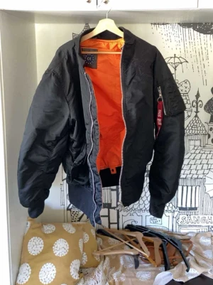 Svart bomberjacka med dödskalle - Säljer en svart bomberjacka med orange innerfoder och broderad dödskalle på ryggen. Jackan har klassisk utility-ficka på ärmen med röd tag och ribbade muddar vid ärmslut och midja. Perfekt för dig som vill ha en edgy och street-inspirerad look.