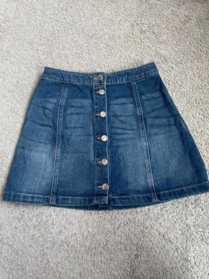 Blå jeanskjol med knappar framtill - Snygg blå jeanskjol i kort modell med raka sömmar och silverfärgade knappar längs hela framsidan. Kjolen har en klassisk denimlook och passar perfekt till en avslappnad stil. Materialet är kraftigt och håller formen bra.