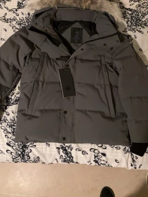 Mörkgrå Canada Goose dunjacka L - Säljer en mörkgrå dunjacka från Canada Goose med stor huva och snygg pälsdetalj. Jackan har klassisk patch på ärmen, dragkedja och knappar framtill samt flera fickor. Perfekt för kalla vinterdagar och riktigt stilren look.