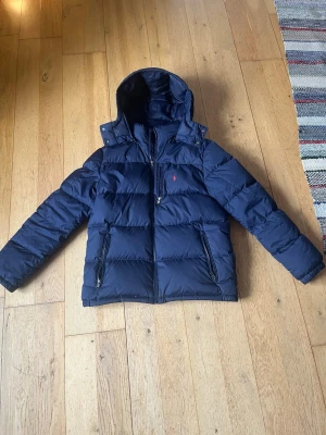 Mörkblå pufferjacka från Polo Ralph Lauren - Snygg mörkblå pufferjacka från Polo Ralph Lauren med huva och dragkedja framtill. Jackan har två sidofickor med dragkedja och en bröstficka med dragkedja. Ikoniska röda Polo-loggan på bröstet. Perfekt för kalla vinterdagar.