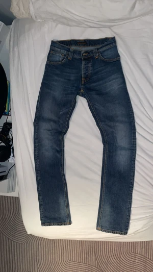 Blå jeans från Nudie Jeans - Säljer ett par klassiska blå jeans från Nudie Jeans med snygg tvätt och raka ben. Jeansen har fem fickor, orangea sömmar och läderpatch bak i midjan. Perfekta för dig som gillar en tidlös och avslappnad stil.
