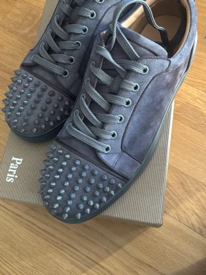 Gråa Christian Louboutin sneakers - Snygga gråa sneakers från Christian Louboutin i mocka med ikoniska nitar på tån. Skorna har matchande grå skosnören och en låg silhuett. Insidan är i beige läder med guldfärgad logga. Perfekta för dig som vill sticka ut med en edgy look.