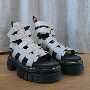 Vita chunky sandaler Dr. Martens - OANVÄND, endast prövade! 🌟🌟🌟  Ricki Gladiator.   Storlek 38 Sulans längd (kant till kant) 25 cm  Pris 1000 kr  De är tillverkade av mjukt Nappa Lux-läder och ger en höjd med en 5-remsdesign, komplett med våra svartagula hälöglor och signaturgula sömmar. Varje par har en gjuten antimikrobiell fotbädd av kork för att hålla fötterna fräscha hela sommaren.  Denna plattform är en lättare konstruktion , Quad Neoteric-sulan är 4,82 cm.