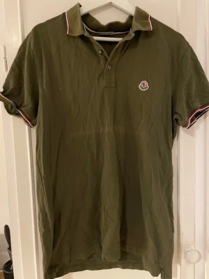 Olivgrön Moncler pikétröja - Säljer en olivgrön pikétröja från Moncler med klassisk krage och korta ärmar. Tröjan har Moncler-logga på bröstet och rödvita detaljer på krage och ärmslut. Perfekt för en avslappnad men snygg stil.