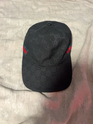 Svart Gucci keps med GG-mönster - Säljer en svart keps från Gucci med klassiskt GG-monogram över hela kepsen. På sidorna finns gröna och röda ränder som ger den en ikonisk look. Kepsen har böjd skärm och är tillverkad i ett slitstarkt tygmaterial. Perfekt för dig som vill ha en stilren accessoar.