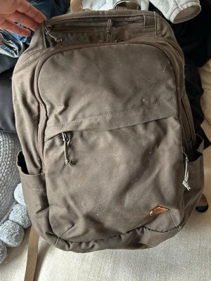Brun ryggsäck från Fjällräven - Säljer en brun ryggsäck från Fjällräven med klassisk rävlogga i läder. Ryggsäcken har flera fack med dragkedja, sidofickor och justerbara axelband. Tillverkad i slitstarkt tyg, perfekt för skola eller utflykter.