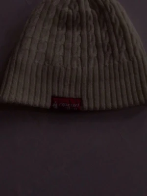 Vit kabelstickad mössa Rip Curl - Säljer en vit kabelstickad mössa från Rip Curl med ribbad kant och ett rött tygmärke framtill. Klassisk modell i mjukt material som håller dig varm och snygg under kalla dagar. Perfekt accessoar för dig som gillar enkel och stilren look.