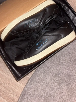  Saint Laurent sneakers - Svarta sneakers från Saint Laurent i slät läder med vit sula och handskriven logga i blått på sidan. Klassisk låg modell med svarta skosnören och minimalistisk design. Perfekta för en clean och stilren look.   