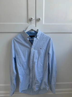 Blårandig skjorta från Tommy Hilfiger - Klassisk blårandig skjorta från Tommy Hilfiger med button-down krage och liten logga broderad på bröstet. Skjortan har långa ärmar, vita knappar och är gjord i mjuk bomull. Perfekt för en clean och stilren look.