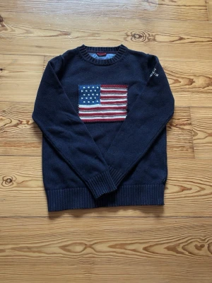 Mörkblå stickad tröja med USA-flagga från gant - Snygg mörkblå stickad tröja med amerikansk flagga i rött, vitt och blått på bröstet. Tröjan har rund halsringning, ribbade muddar vid ärmslut och nederkant samt lång ärm. Perfekt för dig som vill ha en klassisk och cool look.
