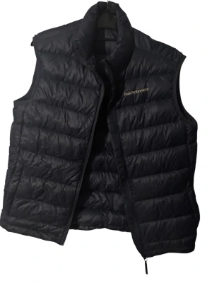 Svart dunväst Peak Performance - Snygg svart dunväst från Peak Performance med quiltad design och hög krage. Västen har dragkedja framtill och diskret logga på bröstet. Tillverkad i lätt och värmande material, perfekt för lager-på-lager och streetwear.