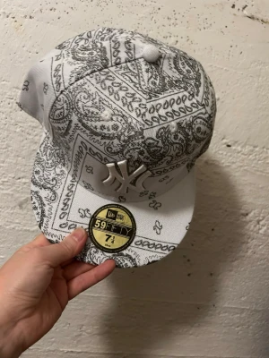 New Era NY Yankees bandanakeps - Säljer en vit New Era 59FIFTY keps med svart bandanamönster och stor NY Yankees-logga i silver framtill. Klassisk flat brim och broderade detaljer. Perfekt för dig som vill sticka ut med streetwear-vibe.